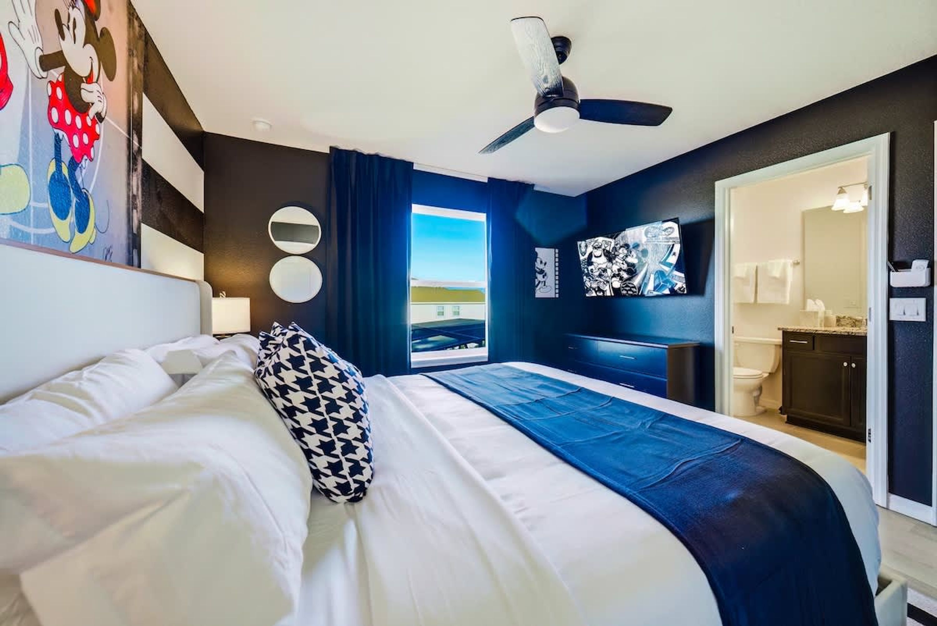 A modern hotel room with a large bed, a ceiling fan, a view of the outdoors, and a bathroom visible through an open door.