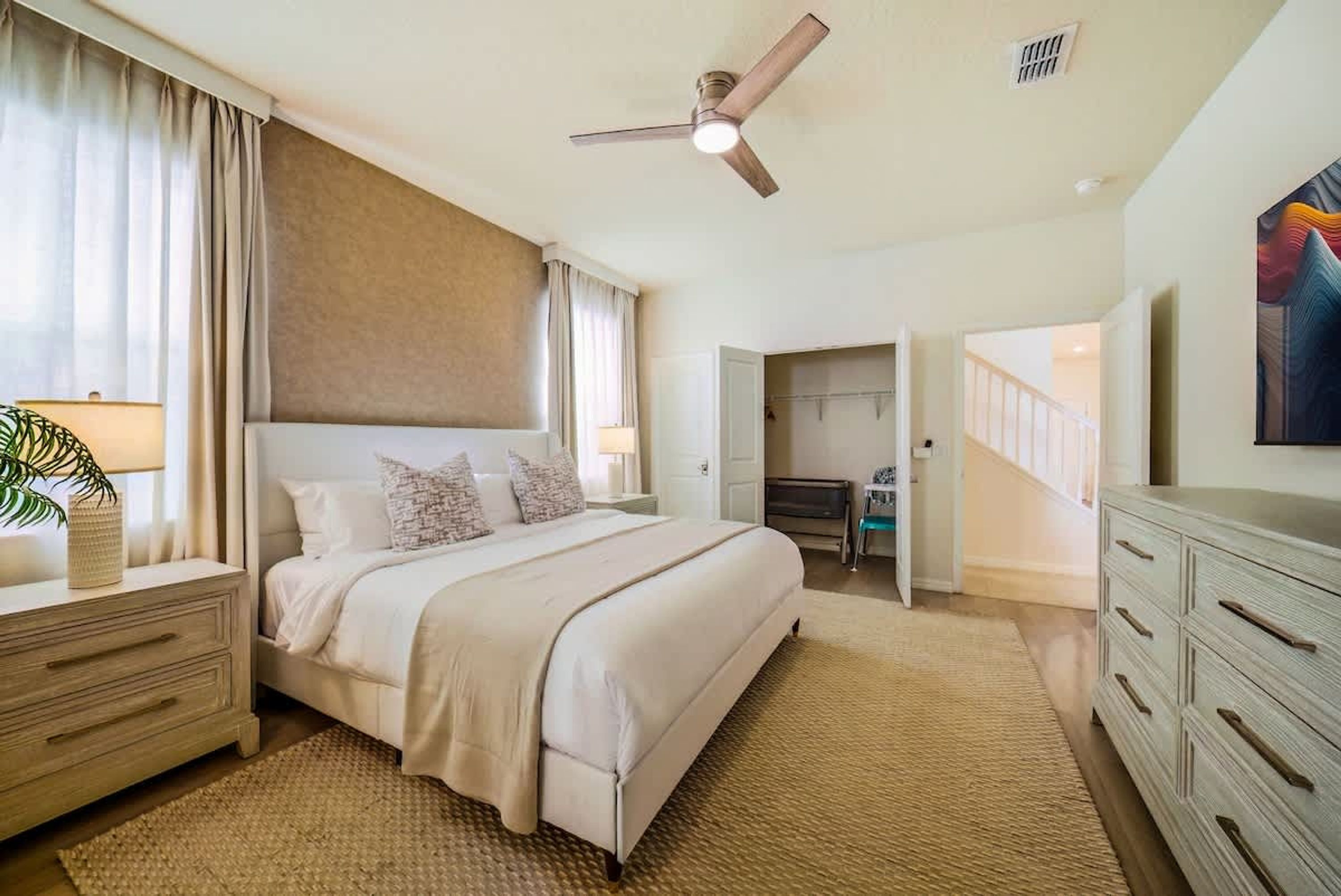 Une chambre à coucher comprenant un lit, des tables de chevet, un ventilateur de plafond, une télévision et une porte donnant sur une salle de bain.