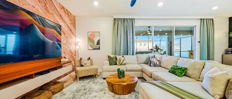 A modern living room with a flat-screen TV, a ceiling fan, a sectional sofa, and a large window.