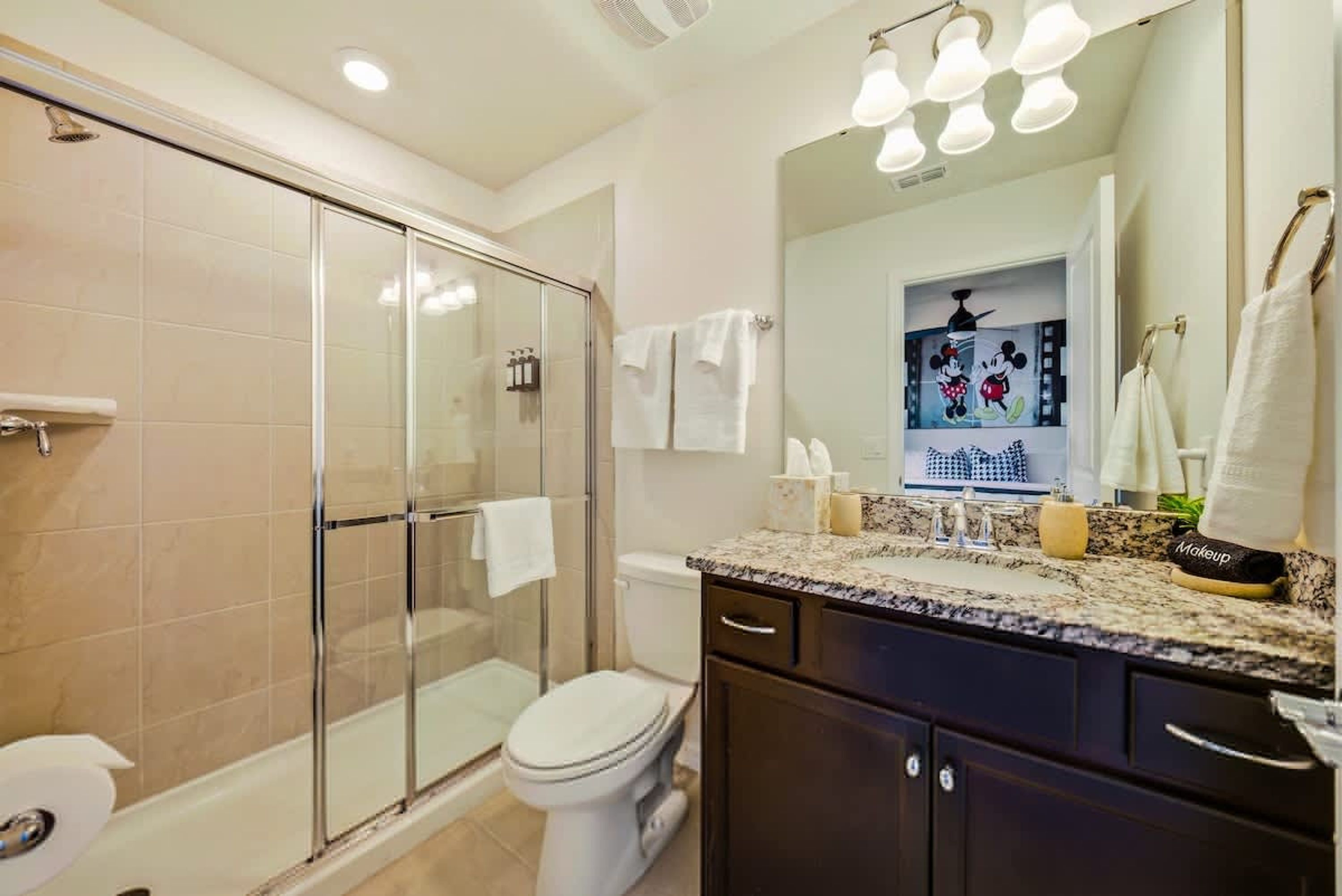 A bathroom with a glass shower enclosure, a toilet, a sink with a vanity, and a mirror with a Mickey Mouse decoration.