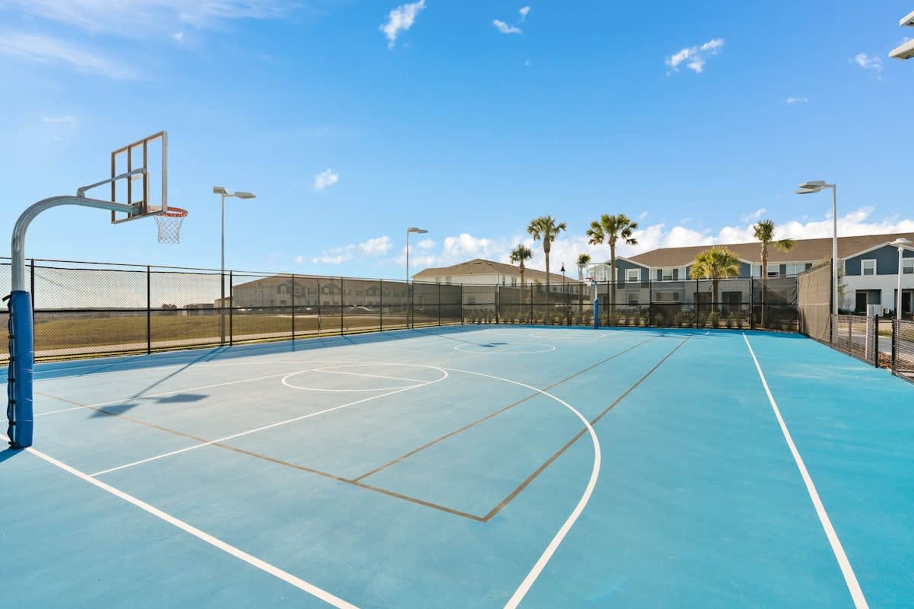 An outdoor basketball court with a blue surface, a basketball hoop, and a chain-link fence.