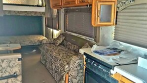 A compact RV kitchen with wooden cabinets, a stove, and a dining area with a patterned sofa. - Peaceful 1-bedroom recreational vehicle with AC in Spring Valley (Spring Valley)