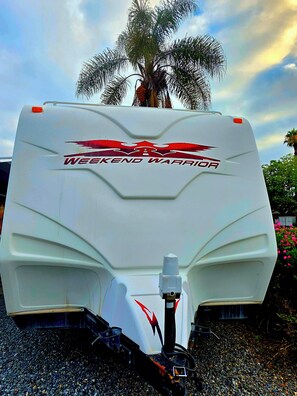 Ein weißer Wohnmobil mit 'Weekend Warrior'-Branding, einer Wasserpumpe und einer Palme im Hintergrund.