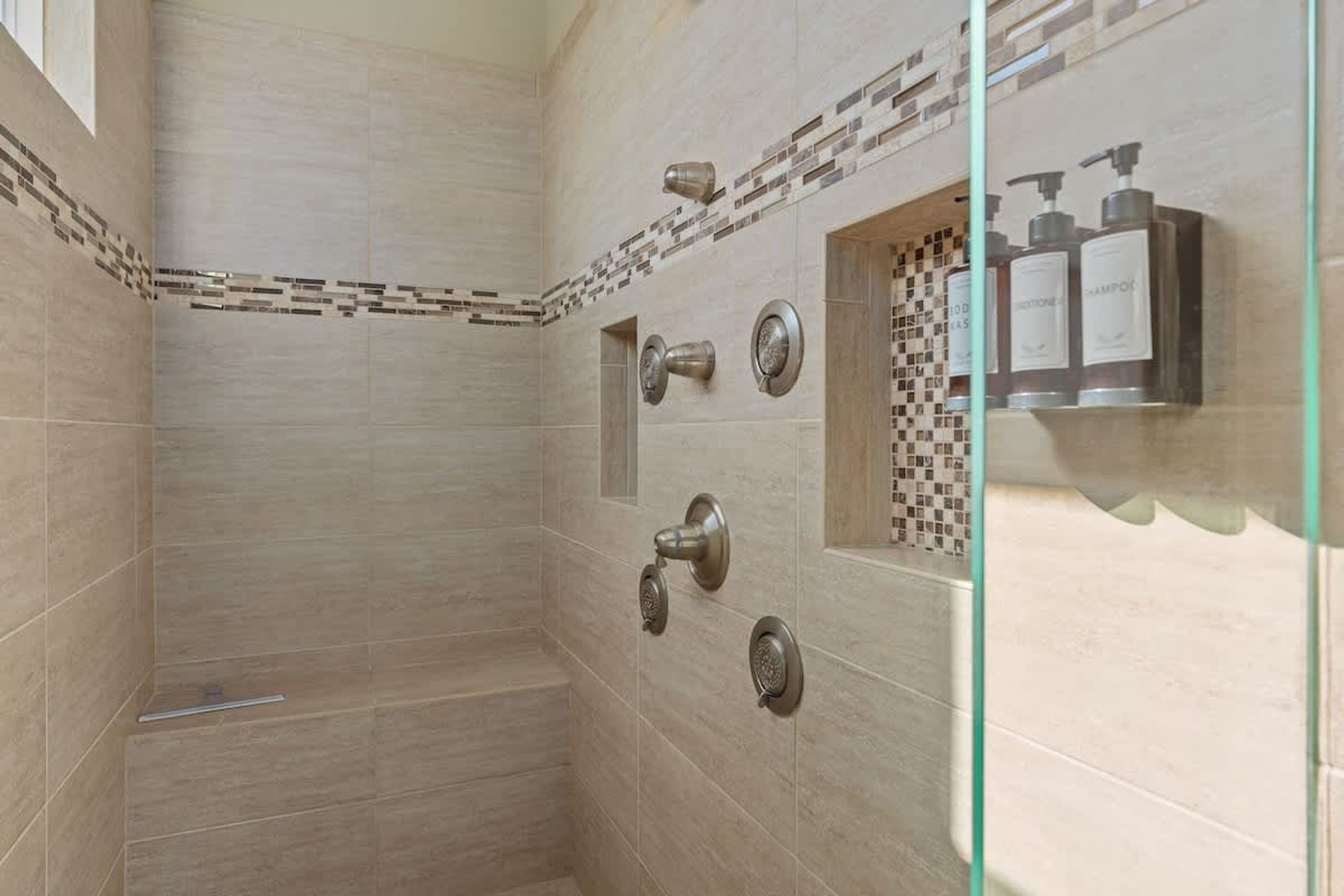 A shower with a glass enclosure, a tiled wall with a checkered pattern, and a shelf with toiletries.