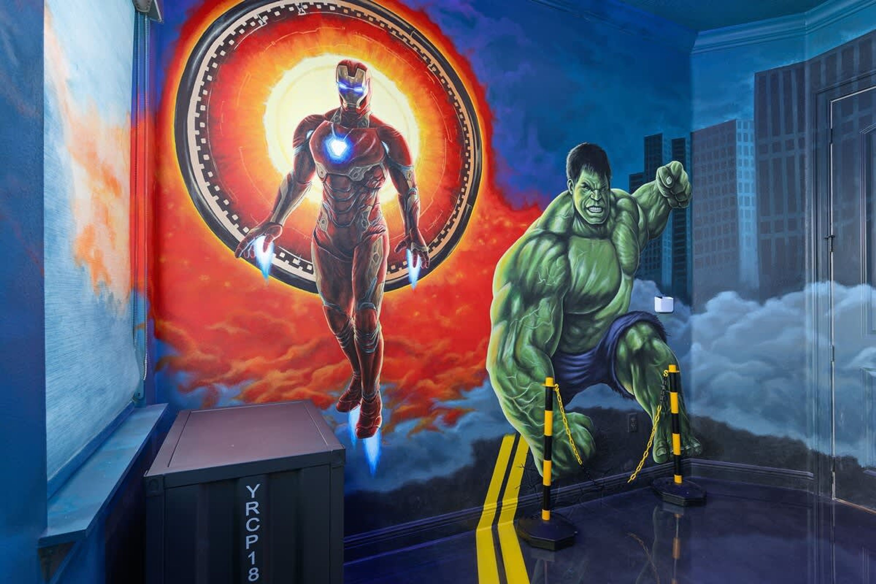 A room with a mural of Iron Man and Hulk, a black box labeled 'YRCP18', and a door to the right.