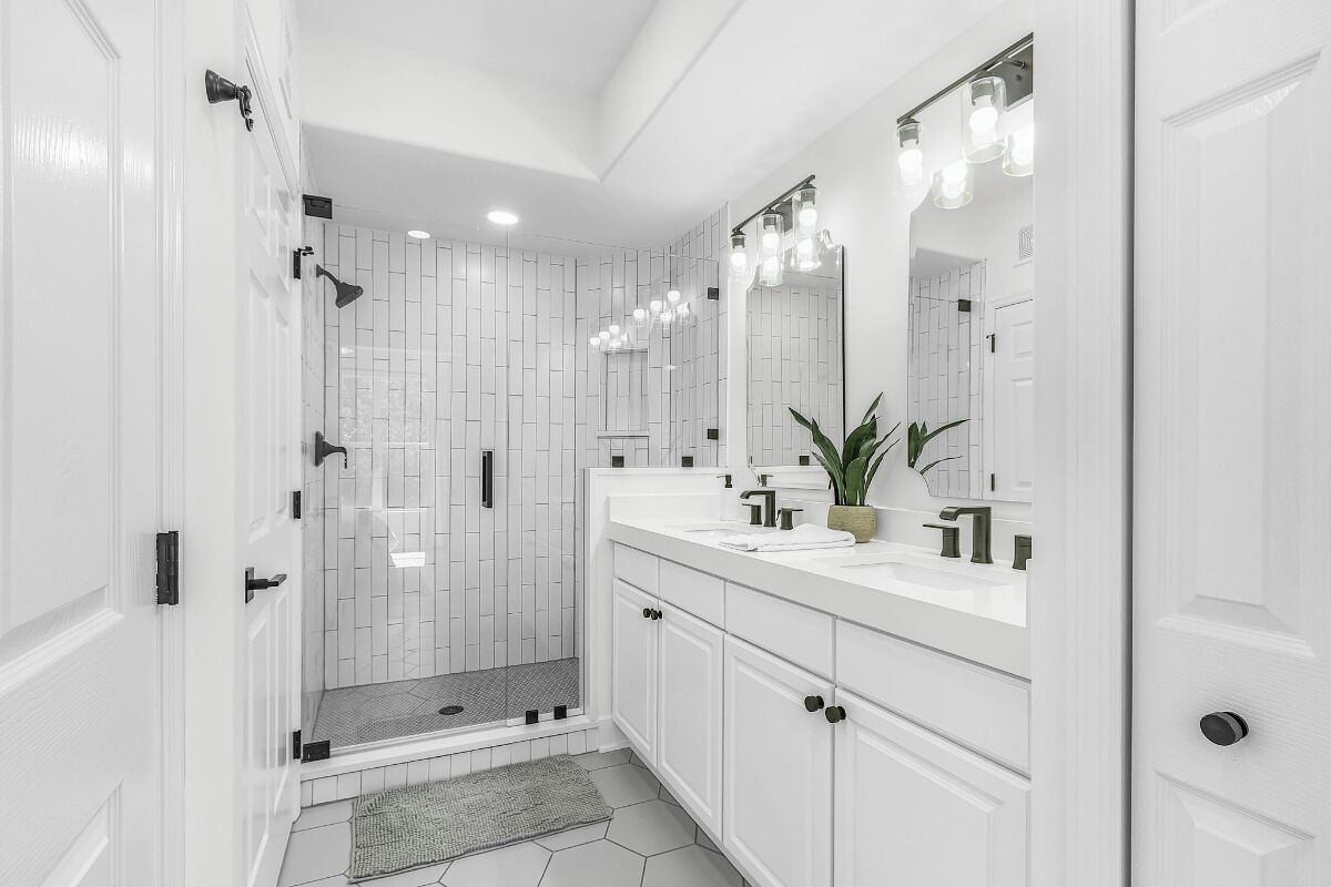 Un baño moderno con azulejos blancos, ducha con puerta de vidrio, tocador con espejo y una planta en maceta.