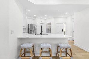 A modern kitchen with white cabinetry, a large island, and three wicker stools. - Completely Remodeled-Fully Furnished Modern 2BR/2BTH-Country Club Membership (Fort Myers)