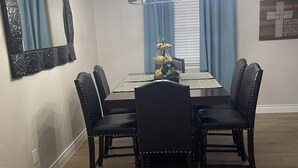 Een eetkamer met een rechthoekige tafel, zes stoelen, een kroonluchter, een spiegel en een raam met gordijnen.