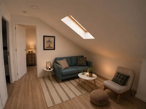 Un dormitori tipus loft acollidor amb un sostre inclinat, un sofà blau, una taula rodona petita, una cadira i una làmpada decorativa.