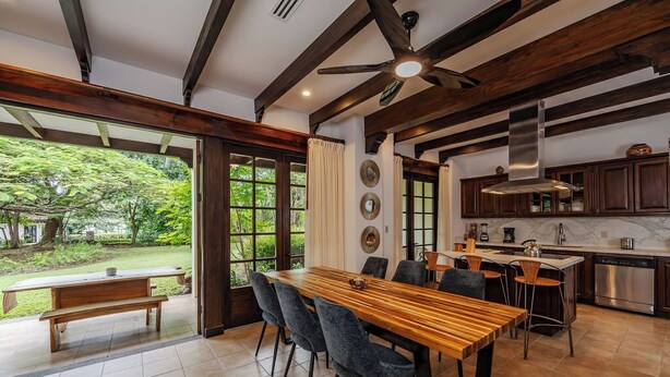 A dining area with a wooden table and chairs, a kitchen with modern appliances, and a view of a garden. - Villa Lima – Relaxing 2-Bedroom Oasis in Hacienda Pinilla (Hacienda Pinilla)