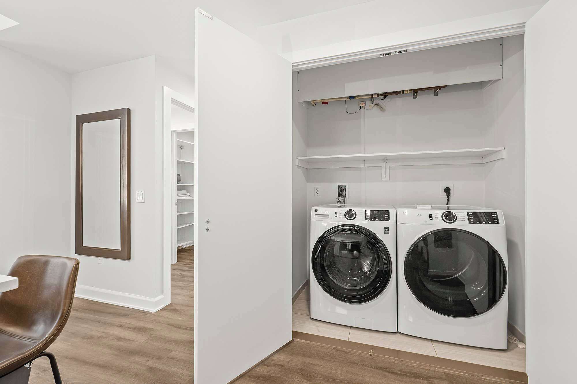 A laundry room with two front-loading washing machines, a wooden chair, and a white shelving unit above the machines.