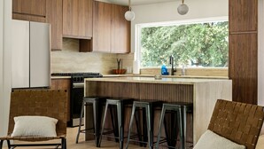 A modern kitchen with wooden cabinets, a central island with bar stools, a window with a view of trees, and a hanging pendant light.