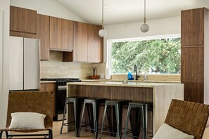A modern kitchen with wooden cabinets, a central island with bar stools, a window with a view of trees, and a hanging pendant light.