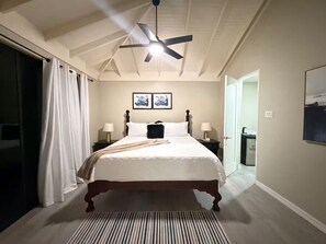 Ein Schlafzimmer mit einem Bett, einem Deckenventilator, zwei Nachttischen mit Lampen, einer Tür und einem gerahmten Bild an der Wand.