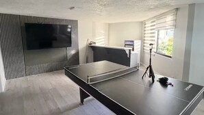 Une chambre moderne équipée d’une table de ping-pong, d’un téléviseur à écran plat et d’une fenêtre avec des stores.