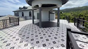 Un espace extérieur abrité doté d’un sol à motifs, d’une structure en forme de gazebo et, au fond, d’un bâtiment avec un toit en tuiles.
