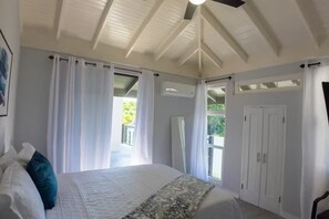 Ein Schlafzimmer mit einem großen Bett, weißen Vorhängen, einem Deckenventilator und Blick auf Grünflächen.