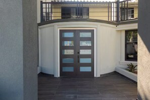 Ein Hauseingang in der Ecke mit einer dunklen Holztür, ein Balkon mit Geländer und ein Fenster mit Blick nach draußen.