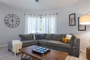 A modern living room with a sectional sofa, a wooden coffee table, a large wall clock, and sheer curtains. - 2 Bedroom Townhouses, Ironshore, Montego Bay (Montego Bay)