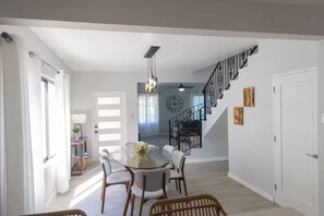 A dining area with a glass table, white chairs, and a wicker chair. A staircase with a black railing leads to a second floor. - 2 Bedroom Townhouses, Ironshore, Montego Bay (Montego Bay)