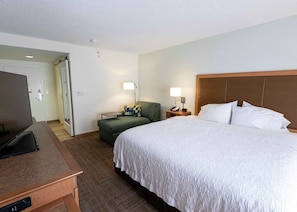 A hotel room with a bed, a desk with a laptop, a green armchair, a nightstand with a lamp, and a television. - Ideal Guestroom for Short Stays–Conveniently Located Near Boca Attractions (Boca Raton)