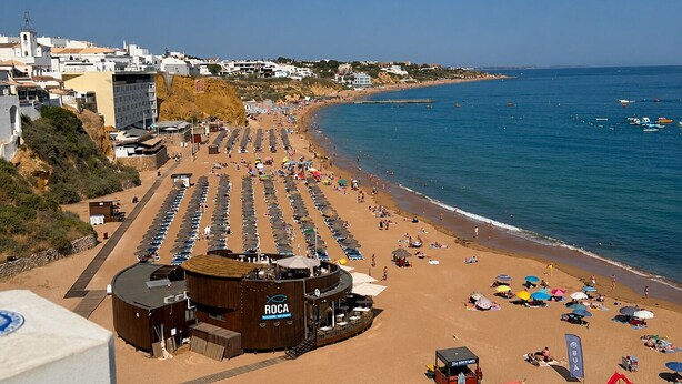 A beach with rows of sun loungers, a wooden building with 'ROCA' signage, and a clear view of the sea.
