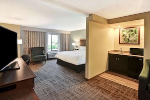 A hotel room with a bed, a desk with a TV, a chair, a window with curtains, a painting, and a kitchenette. - Affordable Unit w/ Breakfast | Quiet & Comfortable Location for Travelers (Boca Raton)