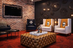 Ein Zimmer mit Backsteinwand, einem Flachbildfernseher, zwei Sesseln mit Totenkopf-Motiven, einem gepolsterten Hocker und dekorativen Wandplatten.