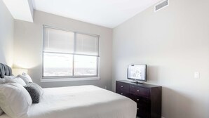 Une chambre à coucher comprenant un lit, une commode, un téléviseur et une fenêtre avec des stores.