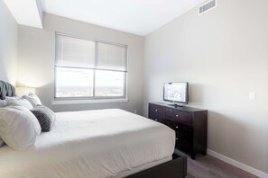 A bedroom with a bed, a dresser, a television, and a window with blinds. - Waterfront Luxury Living in Harbor Point (Stamford, CT)