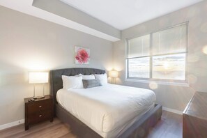 A bedroom with a bed, bedside lamps, a nightstand, a window with blinds, and a painting on the wall. - Waterfront Luxury Living in Harbor Point (Stamford, CT)