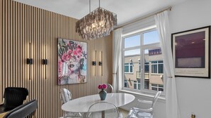 A modern dining area with a round table, clear chairs, a decorative vase with flowers, a large abstract painting, and a view of buildings through a window. - Welcome to Baby Mars: The Ultimate Waterfront Base (Seattle)
