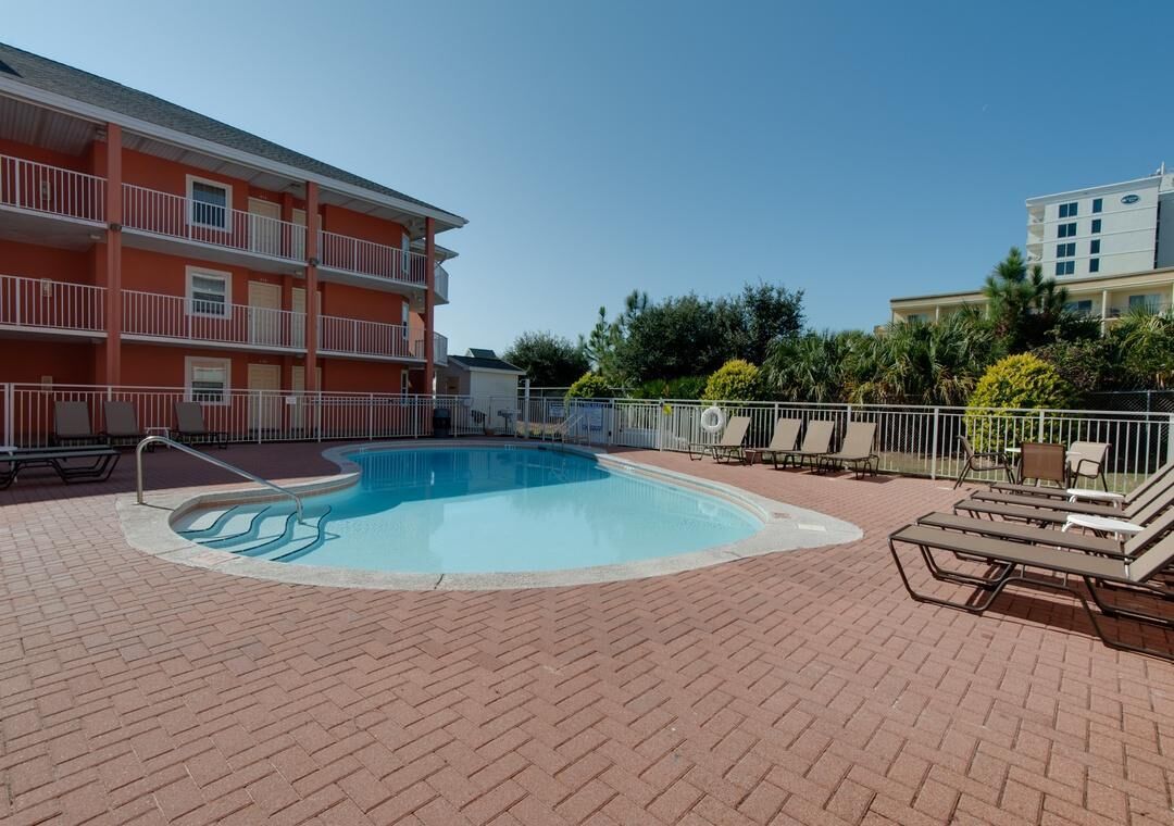 A swimming pool with lounge chairs, a brick patio, and a multi-story building with balconies.
