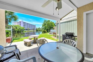 A covered outdoor patio with a glass table, chairs, a grill, and a view of a lake and buildings. - Gulf Highlands 108 - Gulf Breeze Getaway (Panama City Beach)
