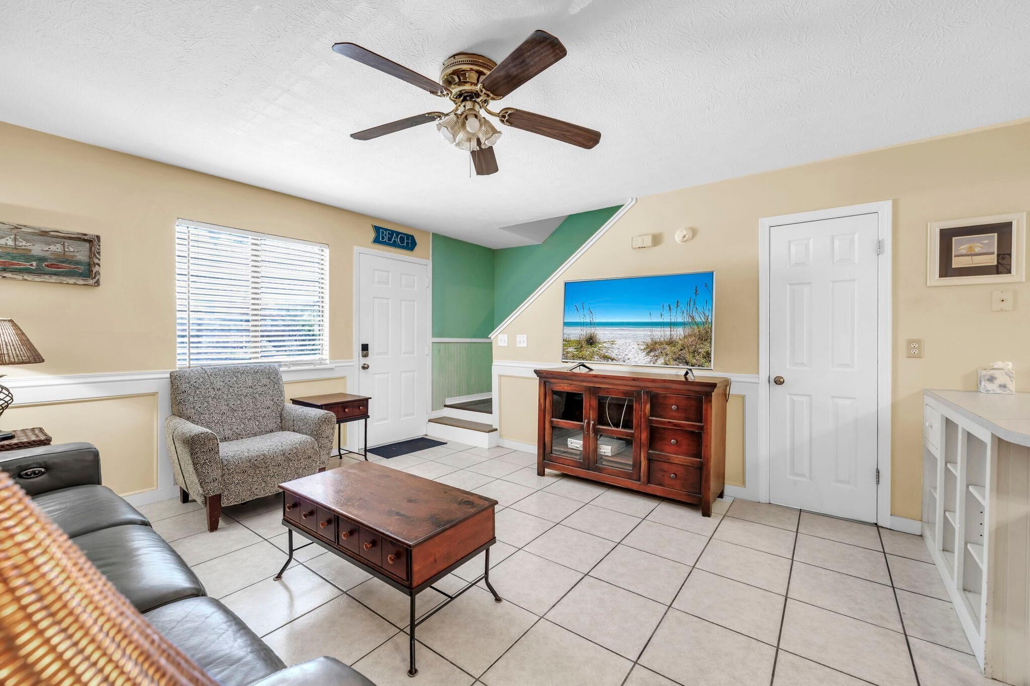 Un salon avec un ventilateur de plafond, un canapé, une table basse, un meuble et un tableau représentant une plage.