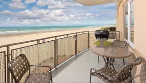 A balcony with a view of the beach, a grill, and outdoor seating.