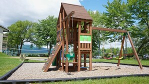 A wooden play structure with a slide and swings, surrounded by gravel and grass, with trees and a building in the background. - O` Bord du Lac Memphrémagog (Magog)