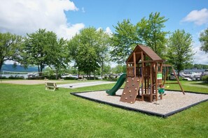 A playground with a climbing wall, slide, and swing set, surrounded by grass and trees. - O` Bord du Lac Memphrémagog (Magog)