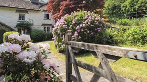 Un bâtiment en pierre doté d’une cheminée, entouré d’une végétation luxuriante et de buissons en fleurs.