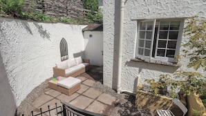 Un bâtiment en pierre avec une façade blanche, une fenêtre, une porte et un petit jardin agrémenté d’un canapé en osier et d’une table.