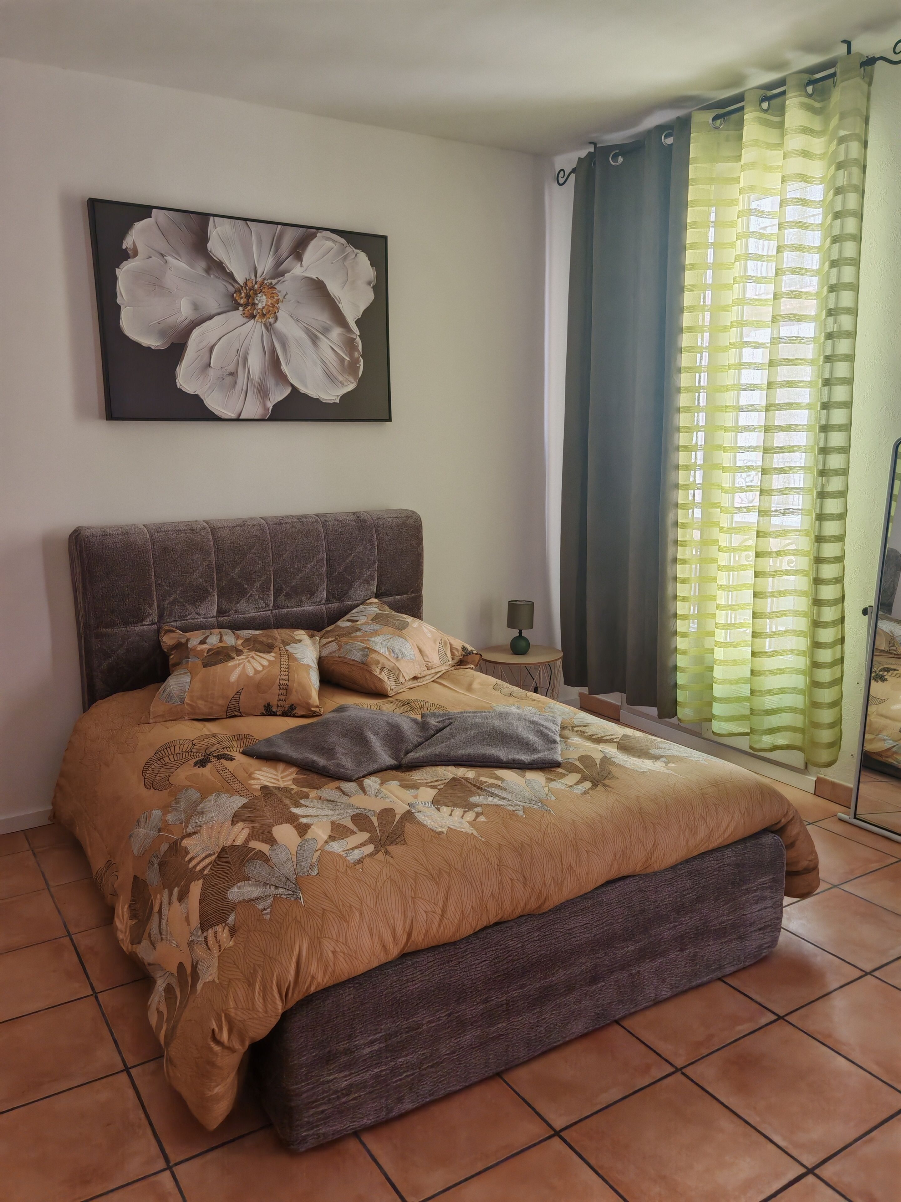 A bedroom with a bed, a headboard, a nightstand, a lamp, a large floral painting, and a window with curtains.