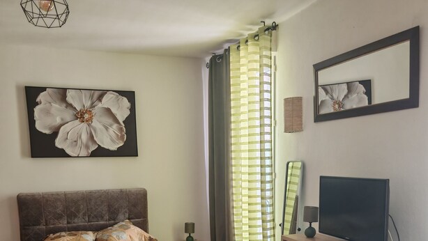 A bedroom with a bed, a television, a desk, a chair, a lamp, and a window with curtains. - A 35-square-meter studio in the heart of downtown, just a stone's throw from all the attractions. (Béziers)