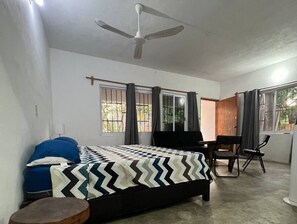 A bedroom with a bed, a desk, and a chair. There is a ceiling fan, windows with curtains, and a view of the outdoors. - Located in the heart of Zicatela (Brisas de Zicatela)