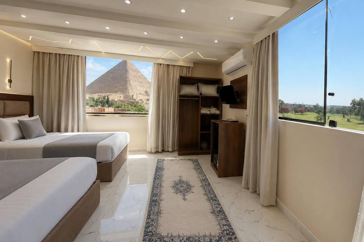 A hotel room with a bed, a desk, a wardrobe, and a large window with a view of a pyramid.