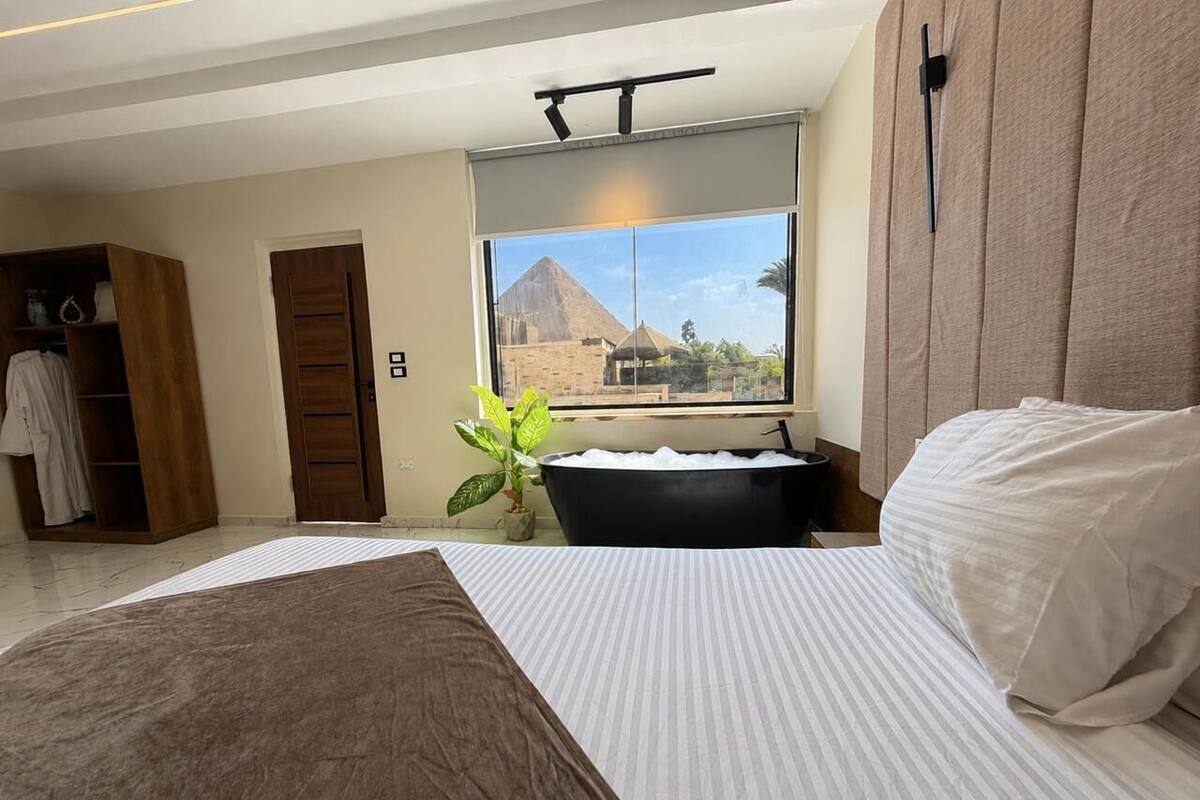 A bedroom with a bed, a bathtub, a window with a view of a pyramid, and a plant.