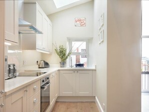 A kitchen with white cabinets, a stove, a sink, a window, and a sign above the window. - Juniper Cottage (Seahouses)