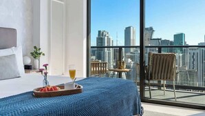 Ein Hotelzimmer mit einem großen Bett, einem Frühstücksbrett mit Obst und einem Getränk sowie einem Balkon mit Blick auf die Stadt.