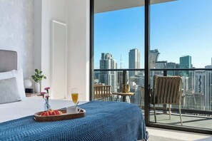 Ein Hotelzimmer mit einem großen Bett, einem Frühstücksbrett mit Obst und einem Getränk sowie einem Balkon mit Blick auf die Stadt.
