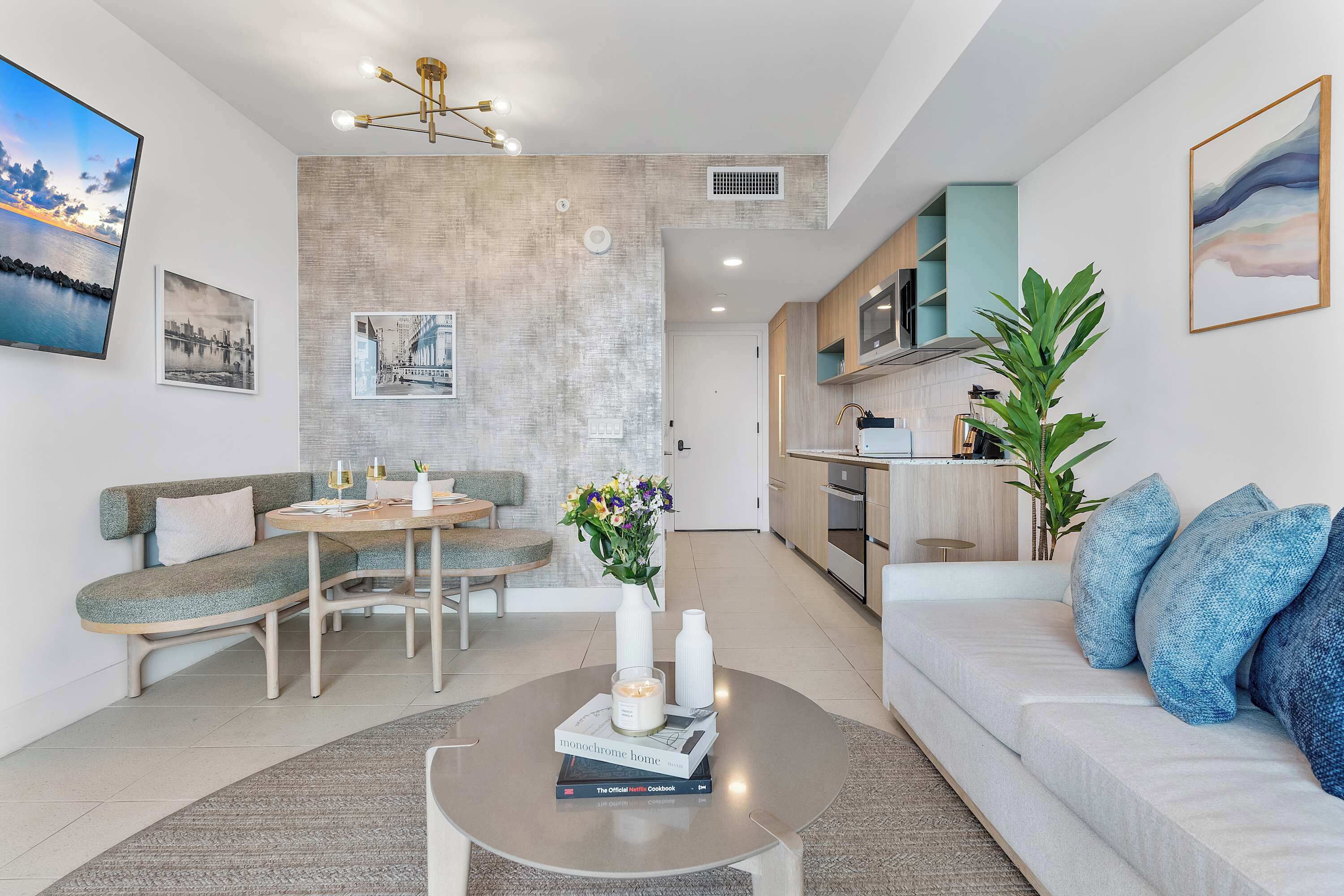 A modern living room with a sofa, a dining table, and a coffee table. There is a kitchen area with a microwave and a plant.