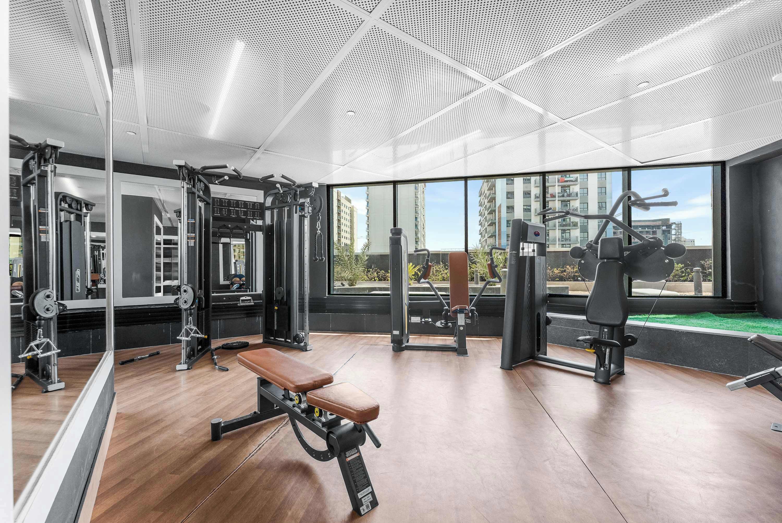 Un gimnasio con equipo de ejercicio, un banco de madera y grandes ventanales con vistas a edificios.